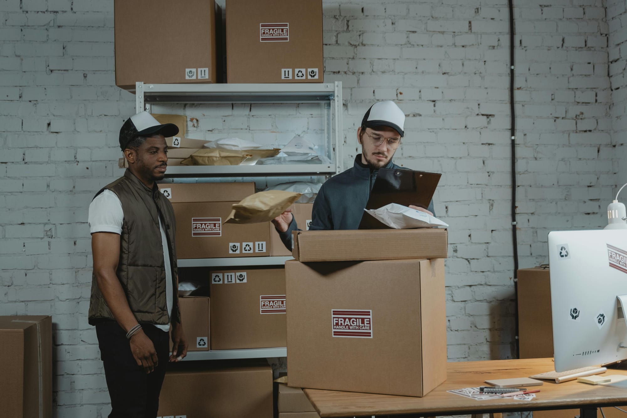 Two men organizing shipping boxes in a warehouse, one checking a clipboard and the other standing near stacked packages.