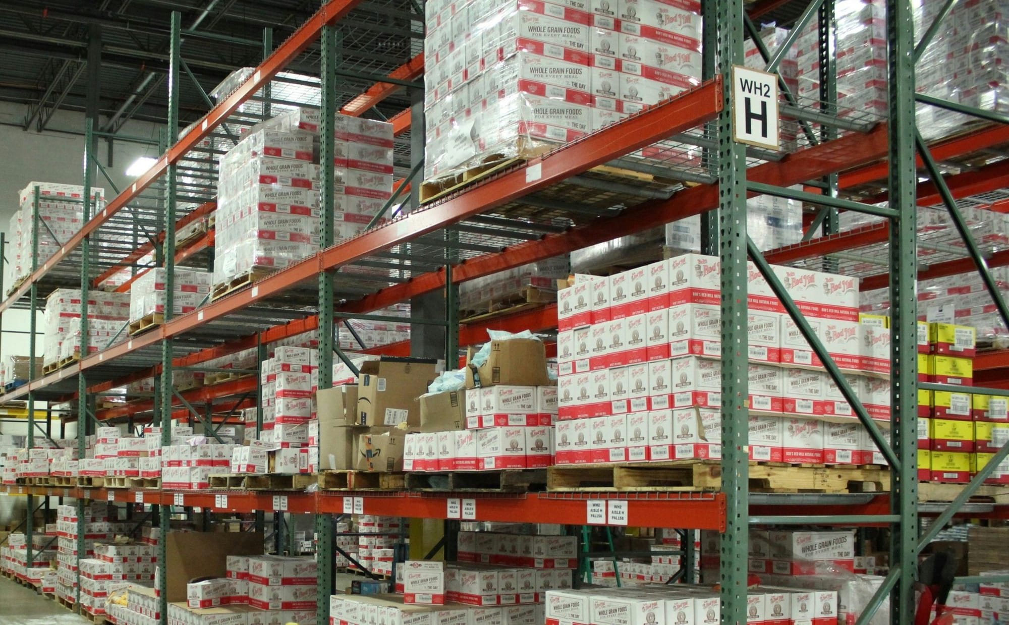 Warehouse racks stacked with pallets of boxed food products, labeled 'Whole Grain Foods,' organized by aisle and section signs.