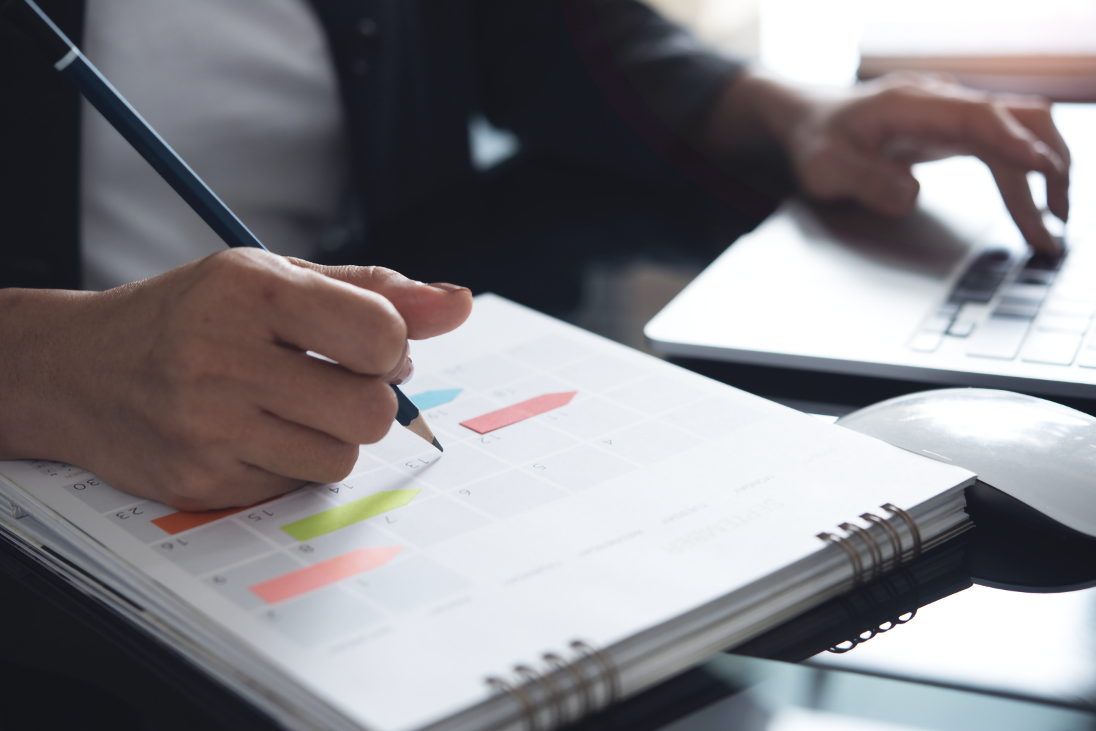 Close-up of a hand marking a shift schedule in a calendar planner, with a laptop in the background.