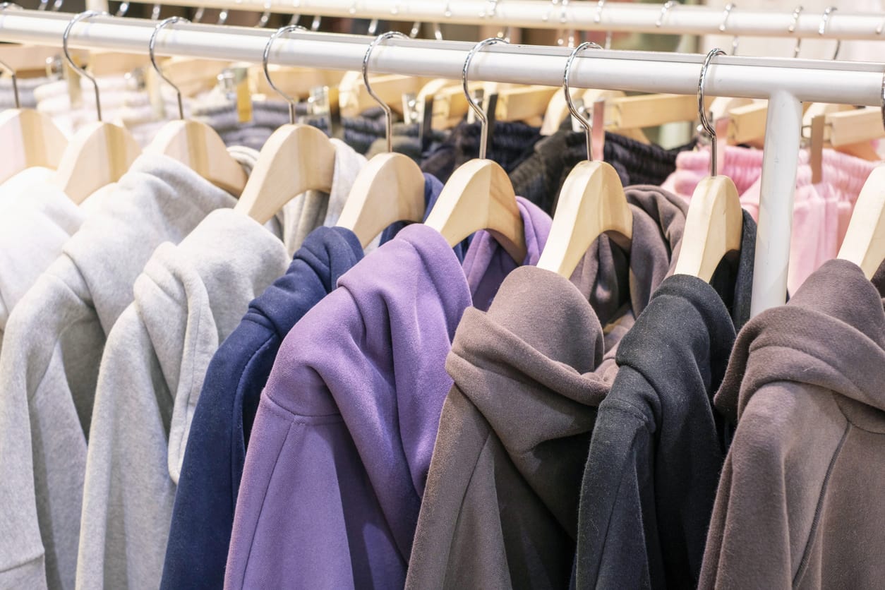 Row of hooded sweatshirts on wooden hangers on a clothing rack in assorted neutral and muted colors.