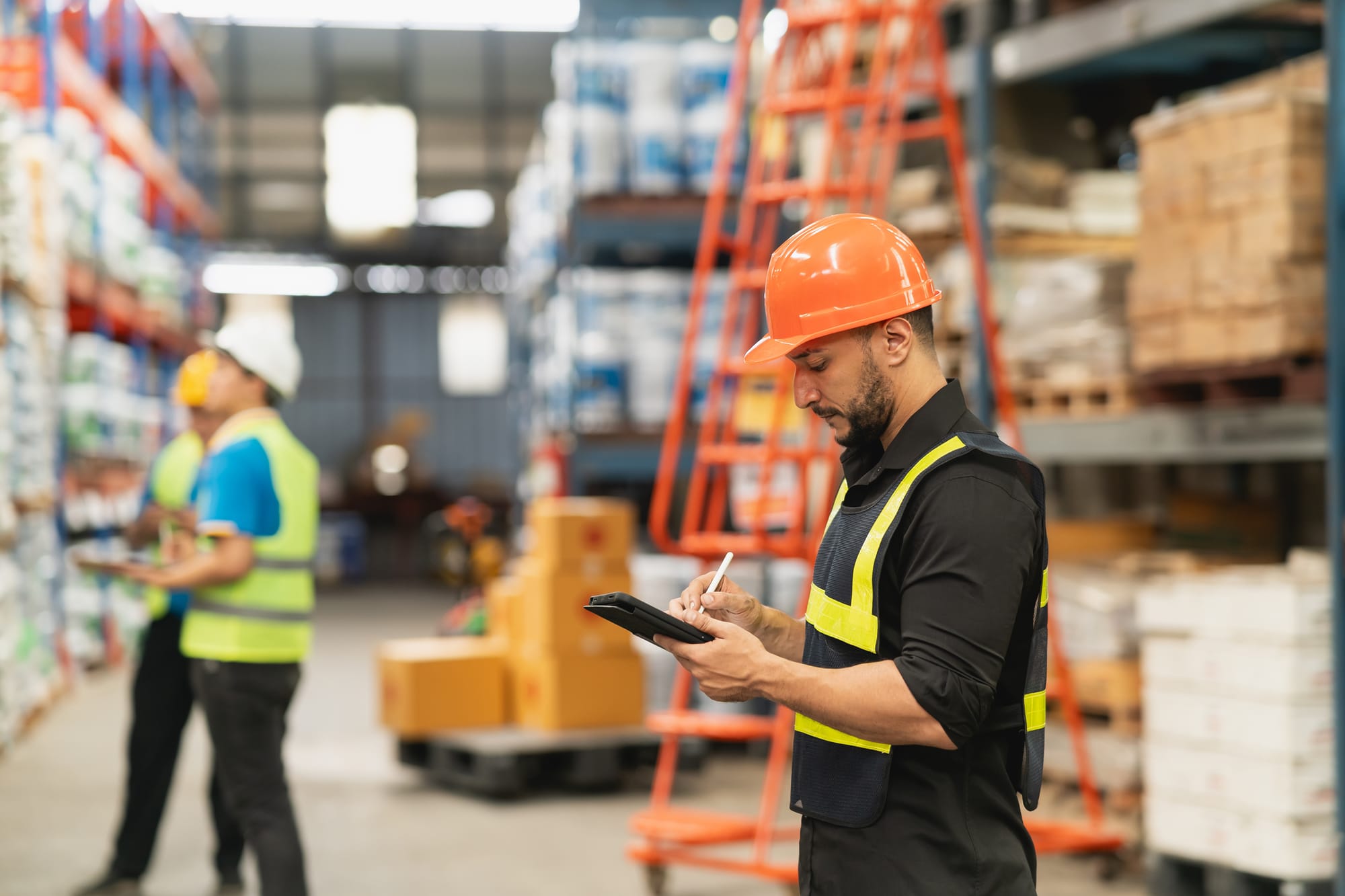 Warehouse employee in a hard hat and safety vest using a digital tablet to check inventory, with boxes and shelves in the background