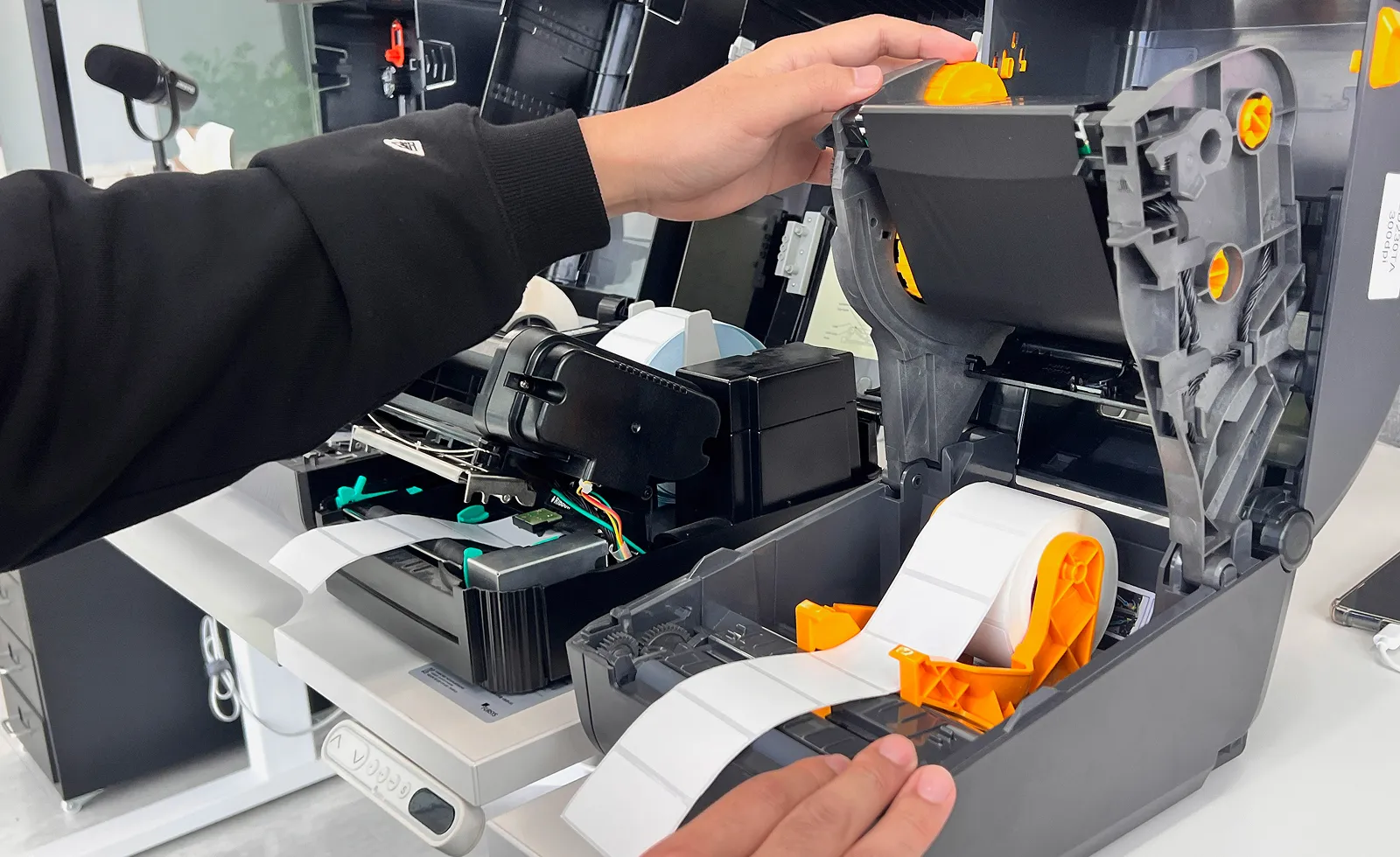 A person loading a label roll into an open Zebra thermal transfer printer, showing how the ribbon mechanism attached to the raised cover creates clear space in the base for easy media access