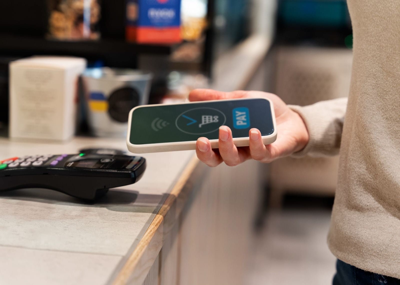 Person holding a smartphone near a contactless payment terminal at a checkout counter, using tap-to-pay.