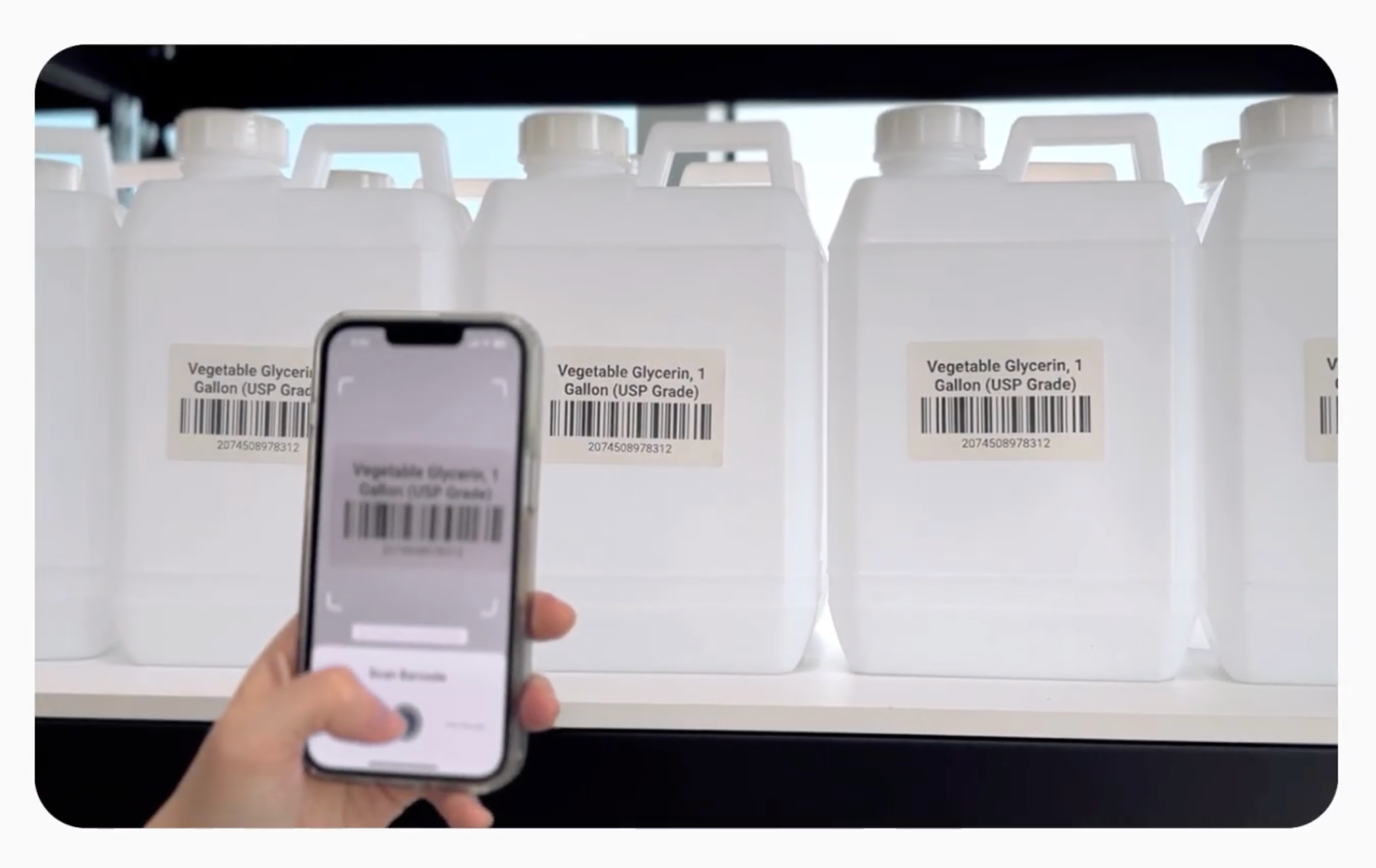 Sebuah tangan memindai label barcode pada galon vegetable glycerin menggunakan aplikasi BoxHero di ponsel