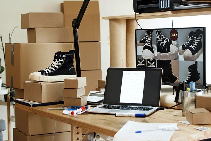 Desk with laptop, stacked shoeboxes, and a single black sneaker on top; product images on a monitor behind.
