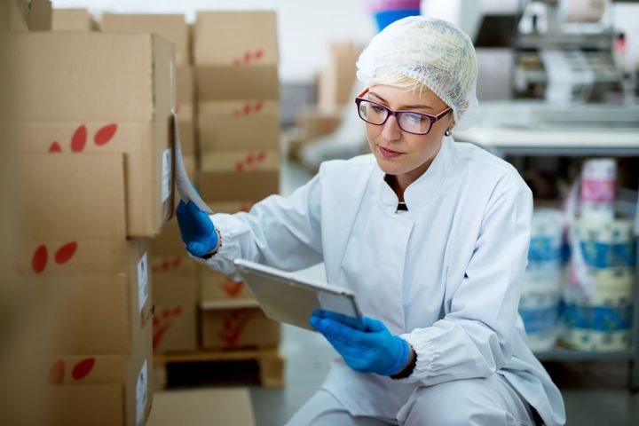 Healthcare worker in a hairnet and gloves checking labels on stacked cardboard boxes with a tablet in a packing area.