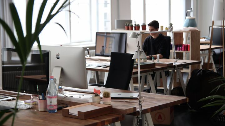 Open office desk with iMacs, plants, and a person working at a table in the background.