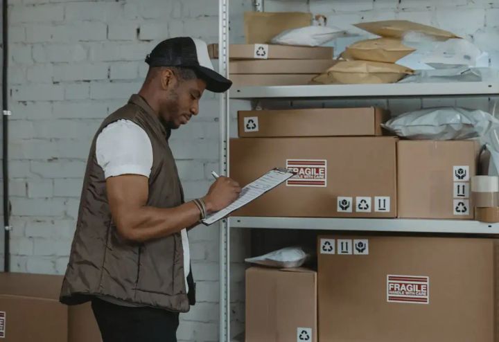 Worker with clipboard checking cardboard boxes on metal shelving; boxes labeled “fragile.”