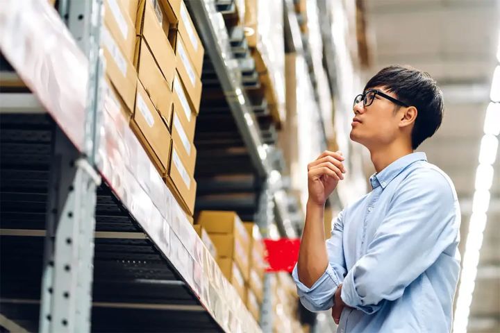 Worker inspecting stacked cardboard boxes on high warehouse shelves, considering inventory or stock locations.