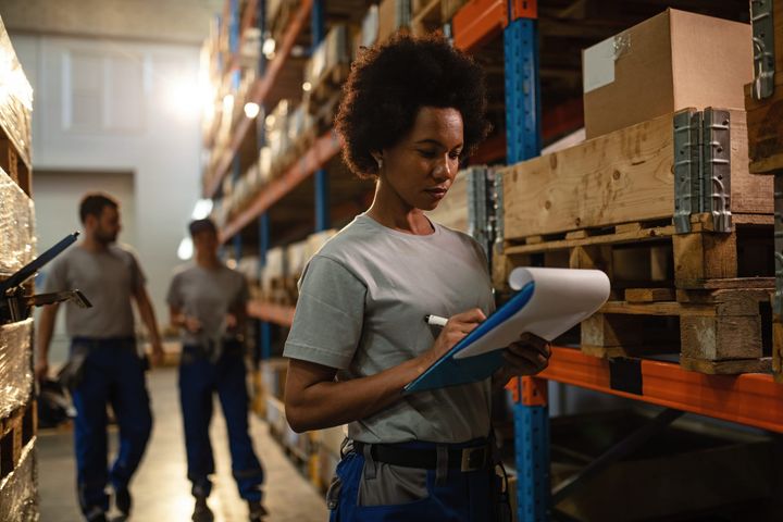 Warehouse worker checking inventory on a clipboard while standing next to pallets and shelves, with two colleagues walking in the background