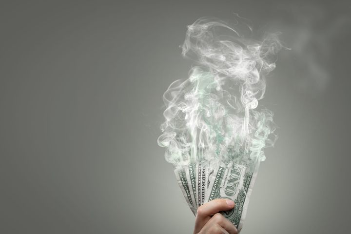 A hand holding several U.S. dollar bills dissolving into the air, resembling smoke.