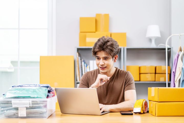 Small business owner working in home office using computer laptop for online sales and inventory management, surrounded by packaged boxes