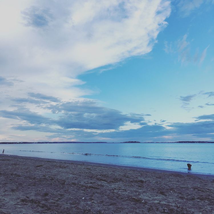 A sandy beach with a few small human figures entering the high tide. The water looks very blue and the sky has some clouds.