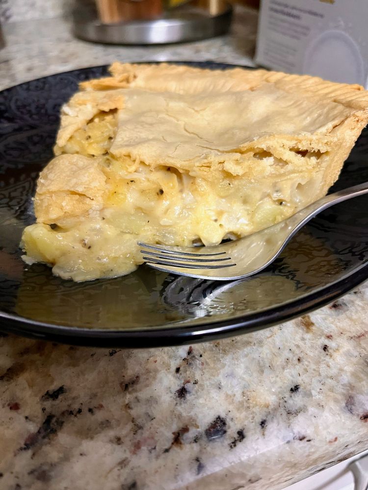 A slice of pie with moist, golden filling speckled by black pepper. It sits on a black ceramic plate with a fork nearby.