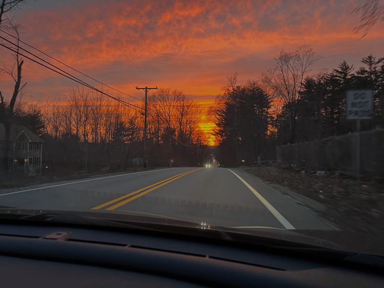 View from a car. Over the road, the Sun is setting in brilliant gold, with pink clouds. Many trees don't yet have leaves.