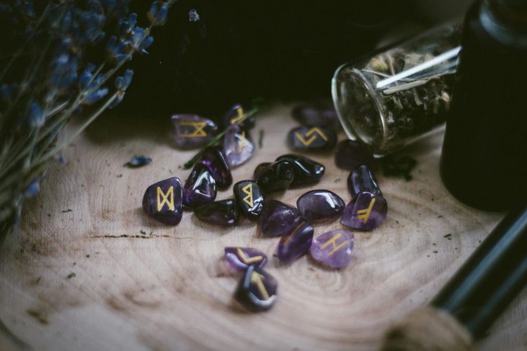 Purple polished gemstones marked with runes in gold paint. They sit on a wooden surface surrounded by herbs.