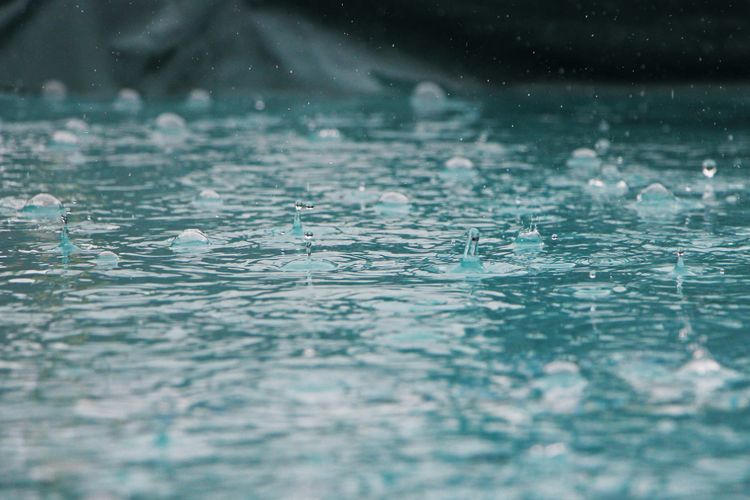 Rain drops landing on a puddle or pool of water. The water reflects a turquoise-blue color, and it ripples.