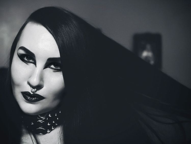 A black and white photo of a woman with straight black hair that she holds out to one side. She has very thick, dramatic eyeliner and glittering black lipstick. Her skin is pale.