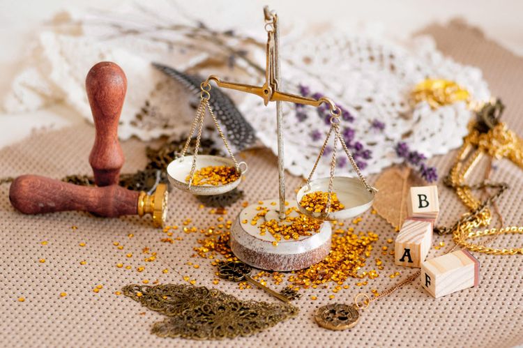 A photographic still life: some ceramic and metal scales loaded with tiny gold discs. Nearby is a seal for wax, some brass trinkets, wooden blocks with black letters, lacework, and flowers.