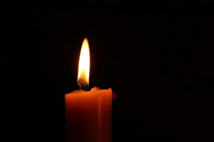 A lit candle that appears orange in the light of its gentle flame. Black background.