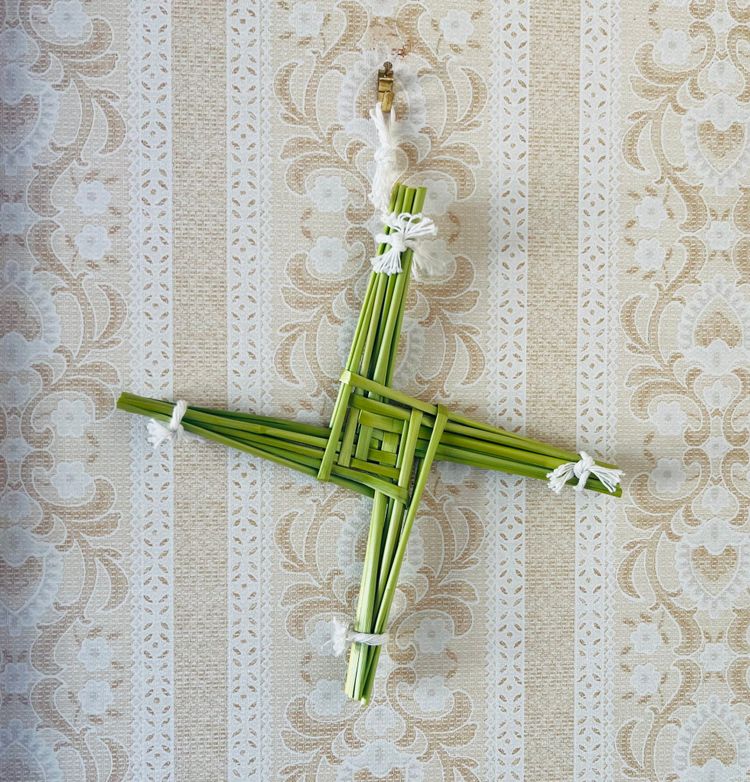 A piece of straw art. It looks like an equilateral cross where the arms seem to emerge from a squared spiral formation in the middle. The straw is green.