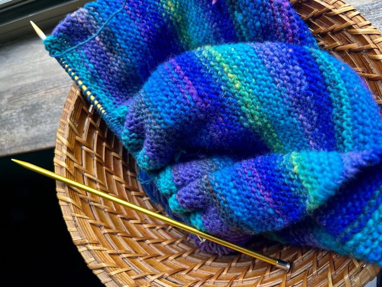 A vibrant blue, indigo, turquoise, and green piece of knitting, on yellow metal needles, in a basket.