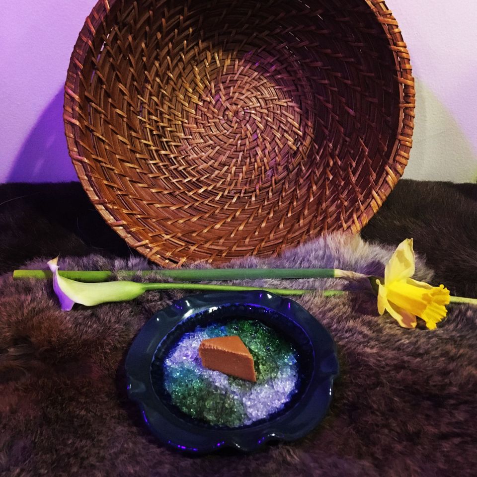 A basket stands upright. Before it is a rabbit pelt with a daffodil, a lily, and a plate containing a piece of Gjetost.