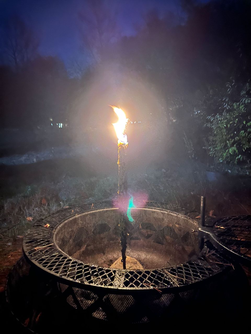 Something that looks like a giant candle is burning on top, in the middle of a metal firepit. It's nighttime beyond.