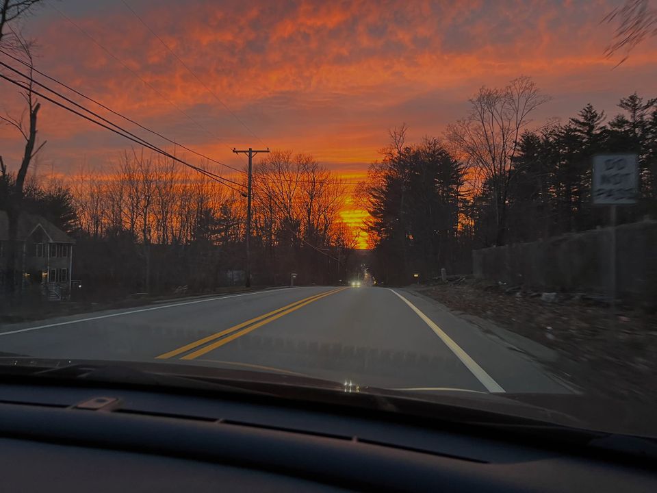 View from a car. Over the road, the Sun is setting in brilliant gold, with pink clouds. Many trees don't yet have leaves.