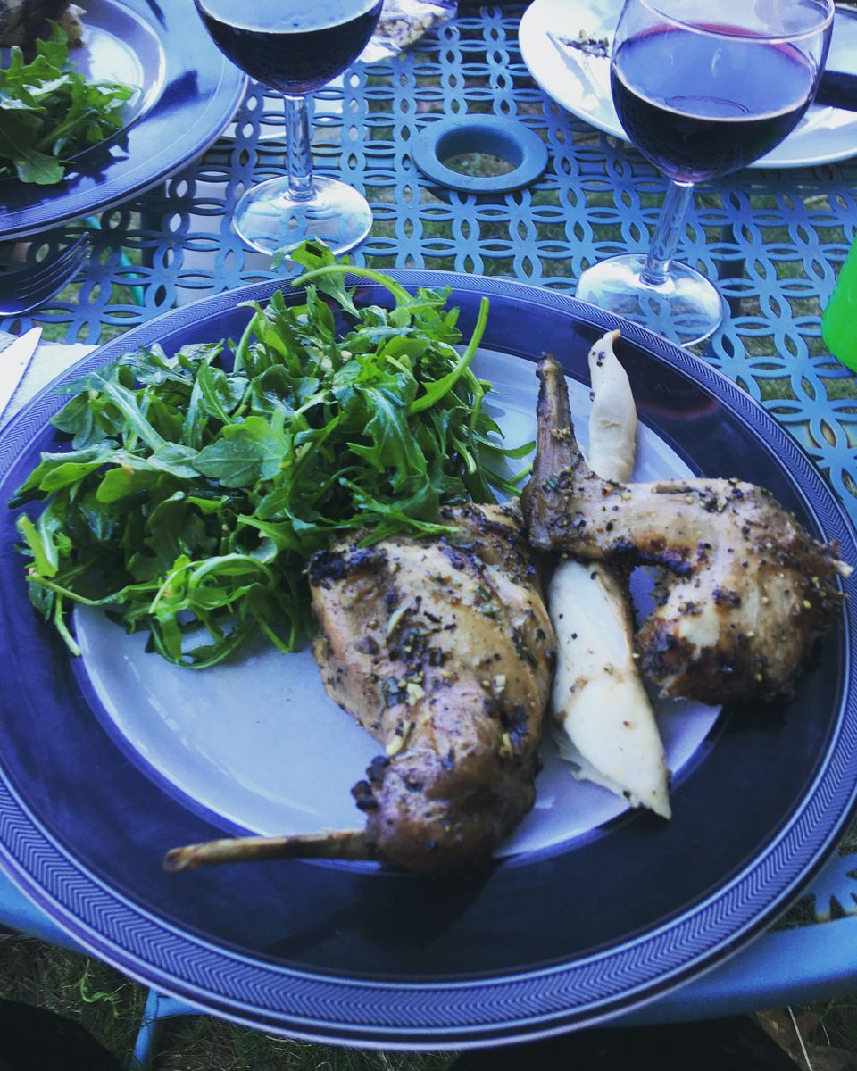 Green arugula and heavily seasoned, cooked rabbit legs and saddle meat on a black and white plate. A glass of wine nearby.
