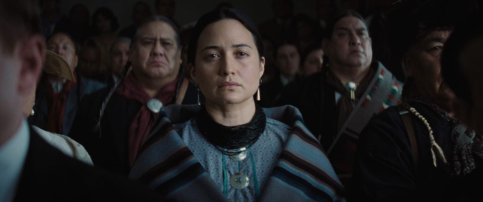 A cinematic shot of several people seated together. The focus is a woman with dark hair, blue clothing, and silver jewelry.