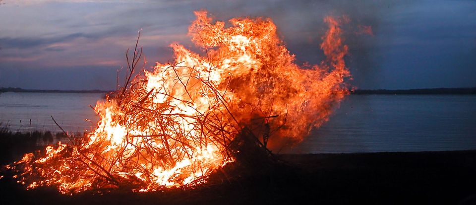A massive bonfire: orange and yellow flames on a large pile of brush. It sits on the shore of a lake at dusk.