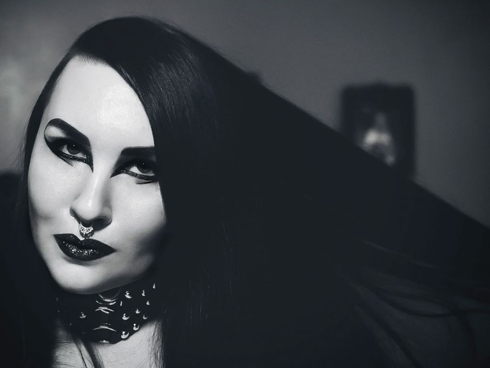 A black and white photo of a woman with straight black hair that she holds out to one side. She has very thick, dramatic eyeliner and glittering black lipstick. Her skin is pale.