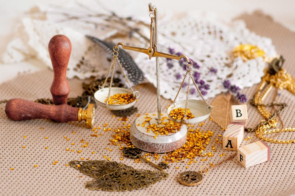 A photographic still life: some ceramic and metal scales loaded with tiny gold discs. Nearby is a seal for wax, some brass trinkets, wooden blocks with black letters, lacework, and flowers.