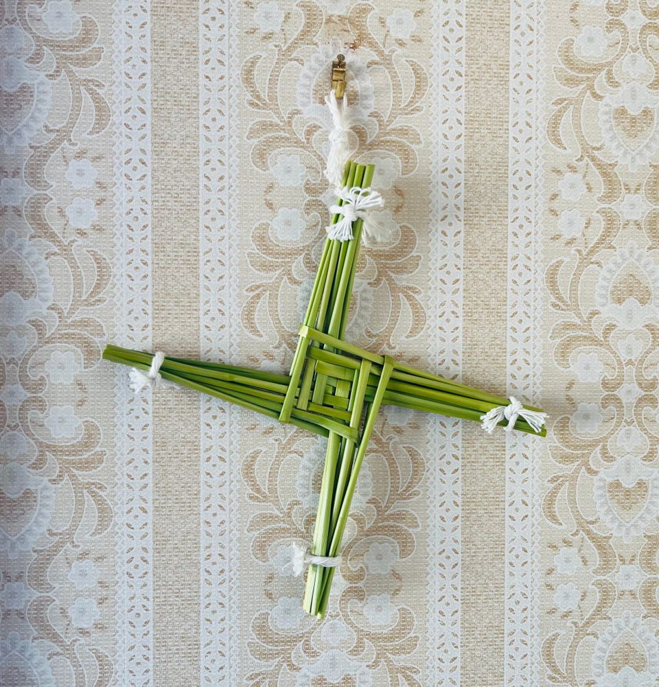 A piece of straw art. It looks like an equilateral cross where the arms seem to emerge from a squared spiral formation in the middle. The straw is green.