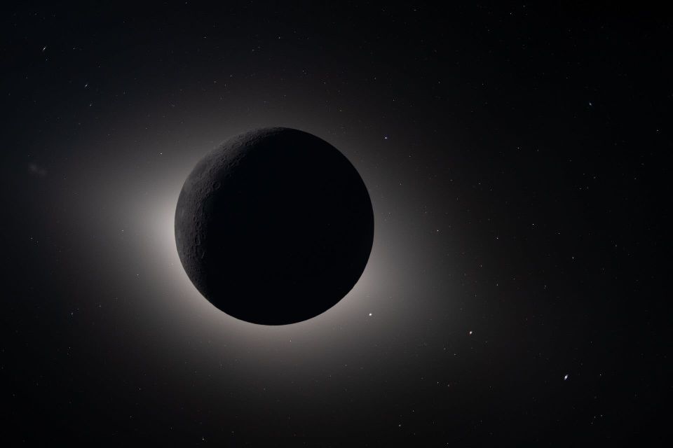 A large grey-black orb, dimly discernible with the features of the lunar surface, covers the Sun's silver corona on a black backdrop speckled by stars and planets.