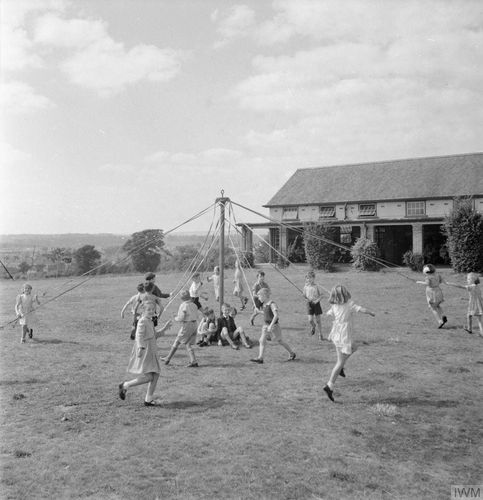 A black & white photograph of young children cavorting with ribbons around a maypole. Those presumably gendered as boys form an inner circle while girls form the outer circle.