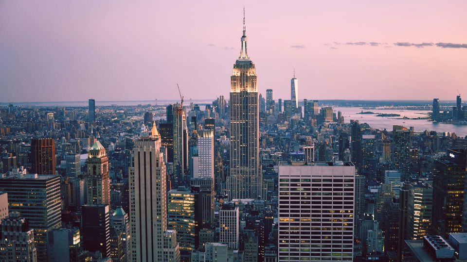 The Manhattan skyline viewed from upon the island itself. Skyscrapers are lit up amid a pink, hazy sundown.