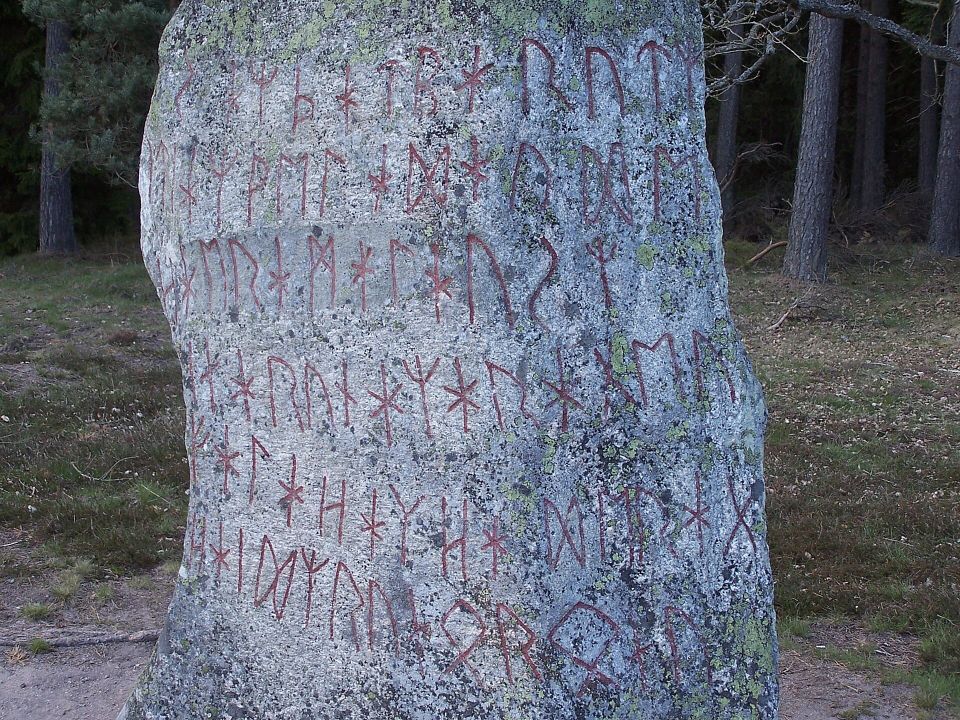Detail of a tall stone inscribed with red runes in a futhark alphabet. Grass and trees are behind it.