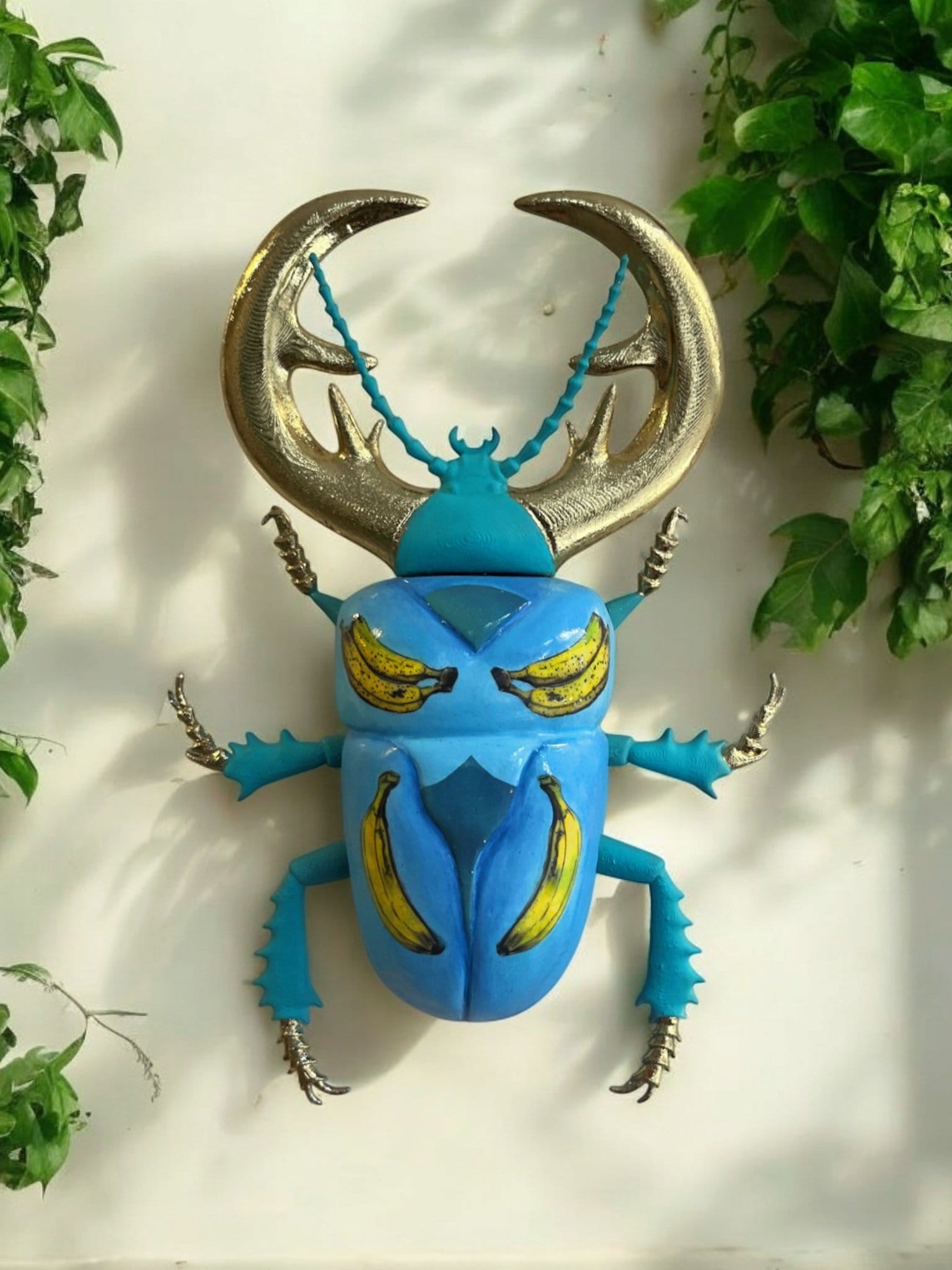 A large 3D-printed beetle mounted on a white wall, with gold antlers, a blue body and yellow bananas painted on its wing cases
