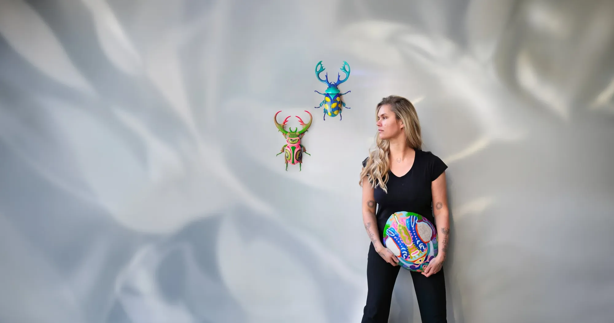 Charlie Moon standing against a pale wall holding a hand-painted round panel, with two colorful 3D-printed beetles mounted above her
