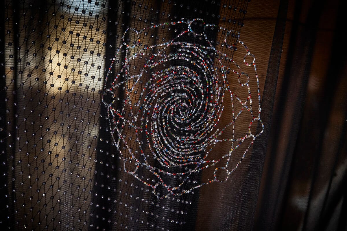 Close detail of a spiral of multicoloured crystals hand-beaded onto black netting, lit from behind