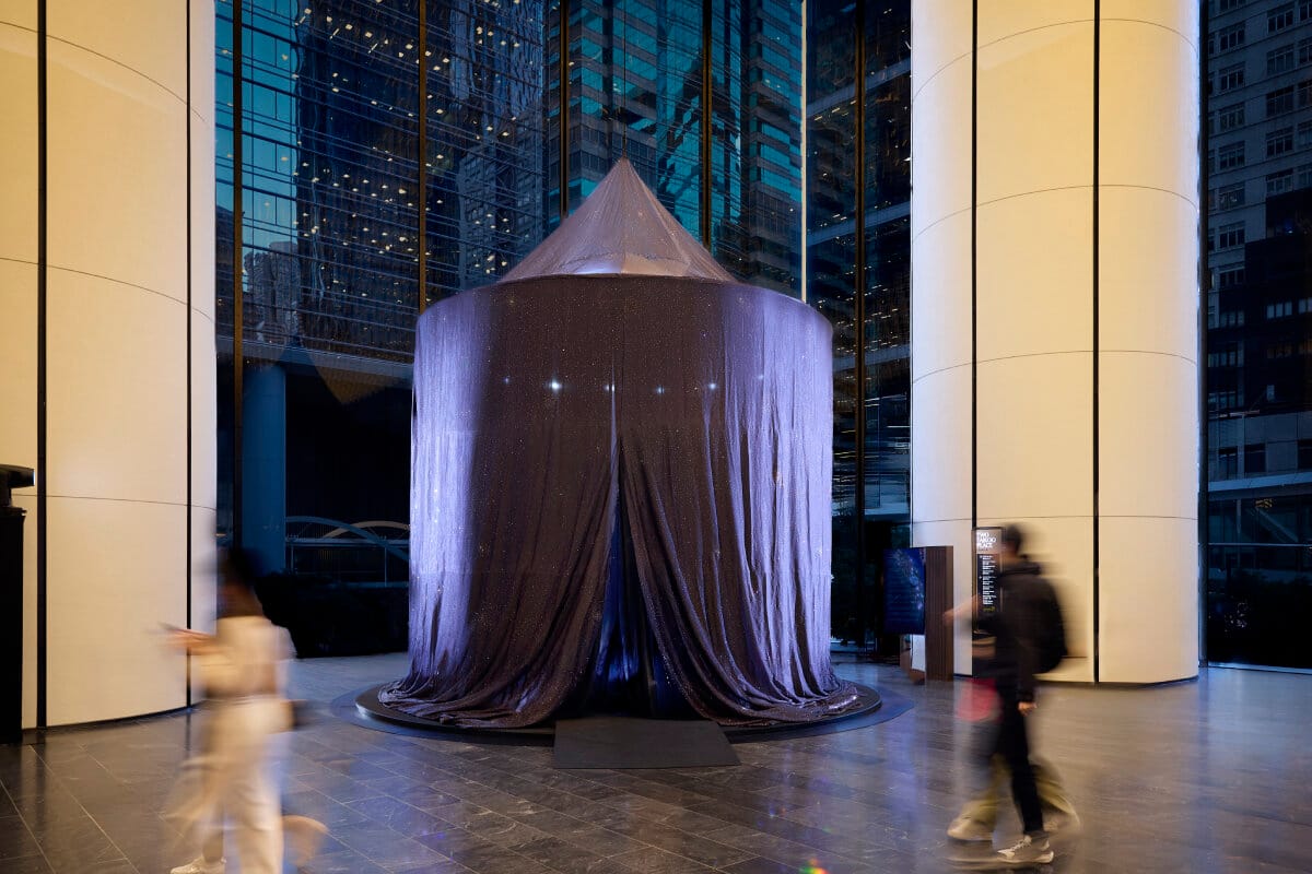 A tall circular tent of dark crystal-flecked fabric standing in a glass office lobby at dusk, its skirt pooling on the floor, with people walking past in motion blur