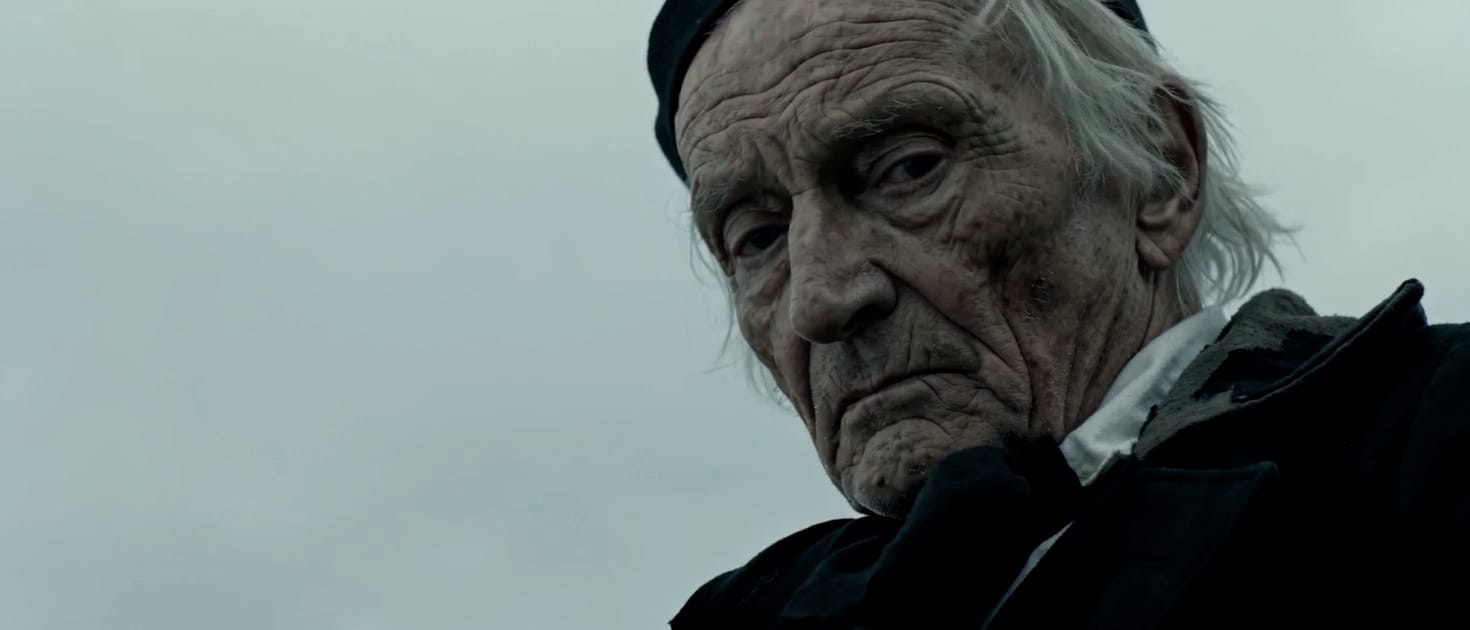 An elderly villager in a black coat against a grey northern sky, a still from Brine
