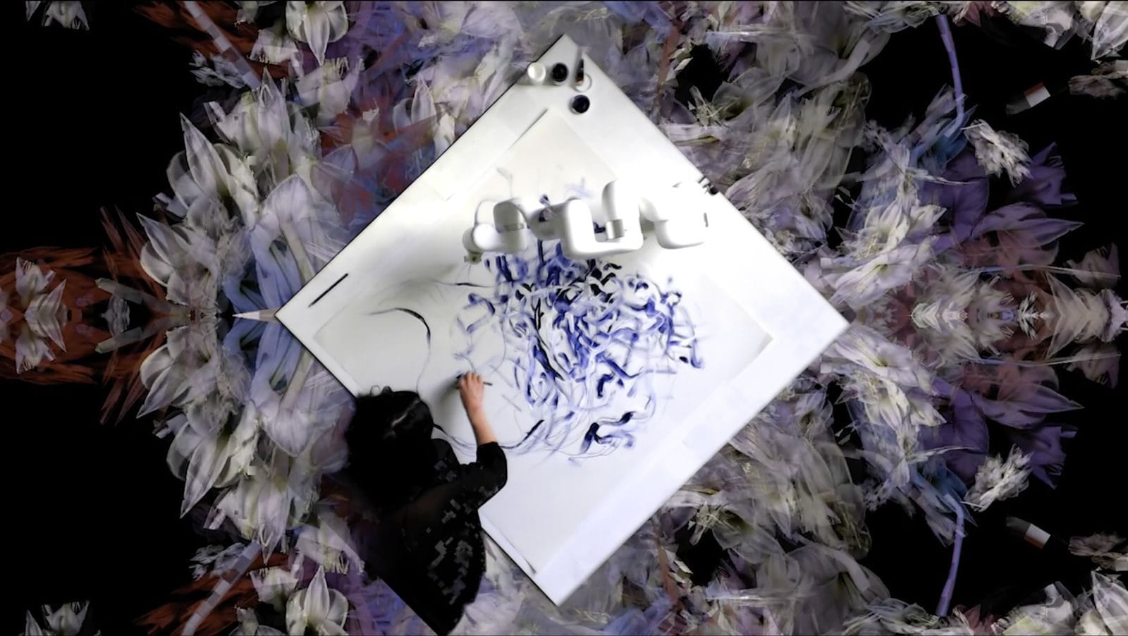 Overhead view of Sougwen Chung drawing on a large white surface alongside robotic units, surrounded by projected imagery