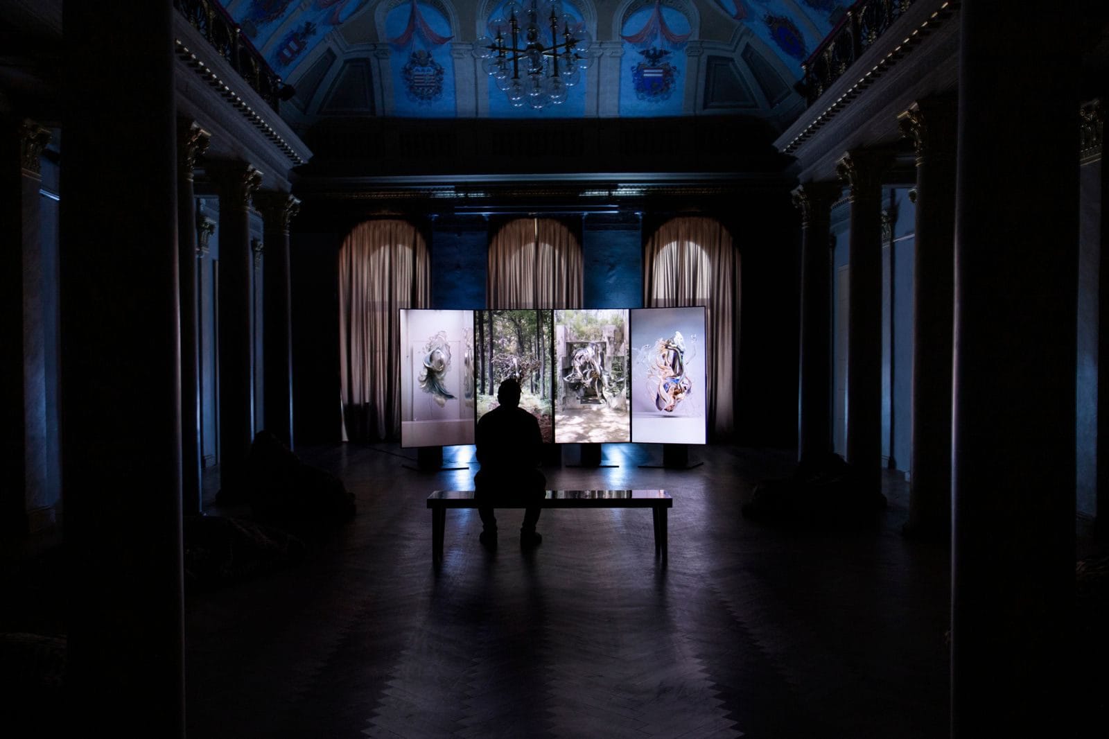 A darkened palazzo gallery with a frescoed ceiling, a single visitor on a bench facing four tall illuminated screens showing swirling forms