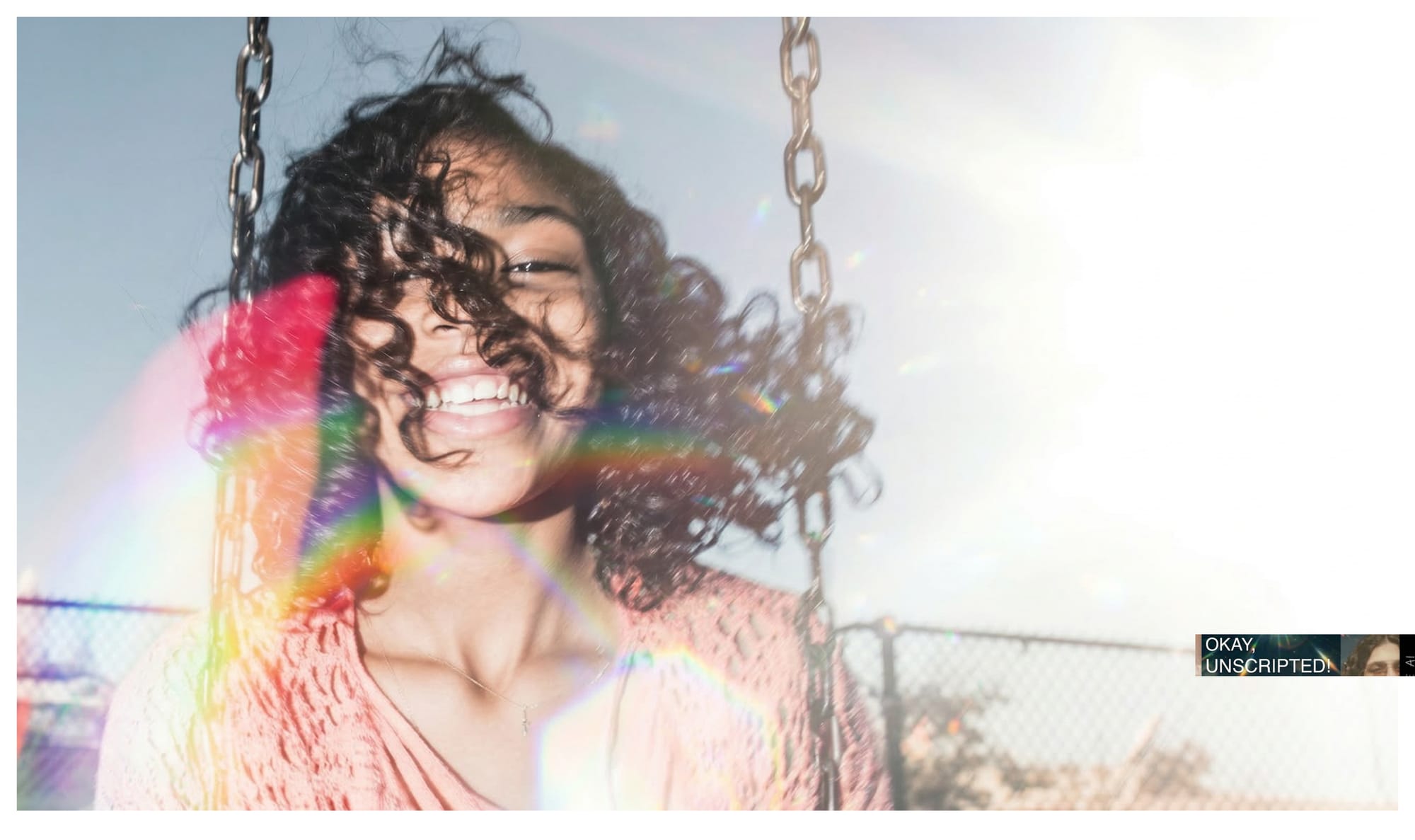 A laughing woman with curly hair on a swing, backlit by bright sun with rainbow lens flare across her face