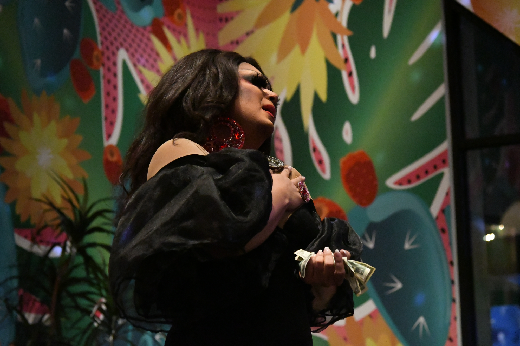 A performer holds cash, wearing large earrings and black attire, in front of a vibrant, colorful wall.