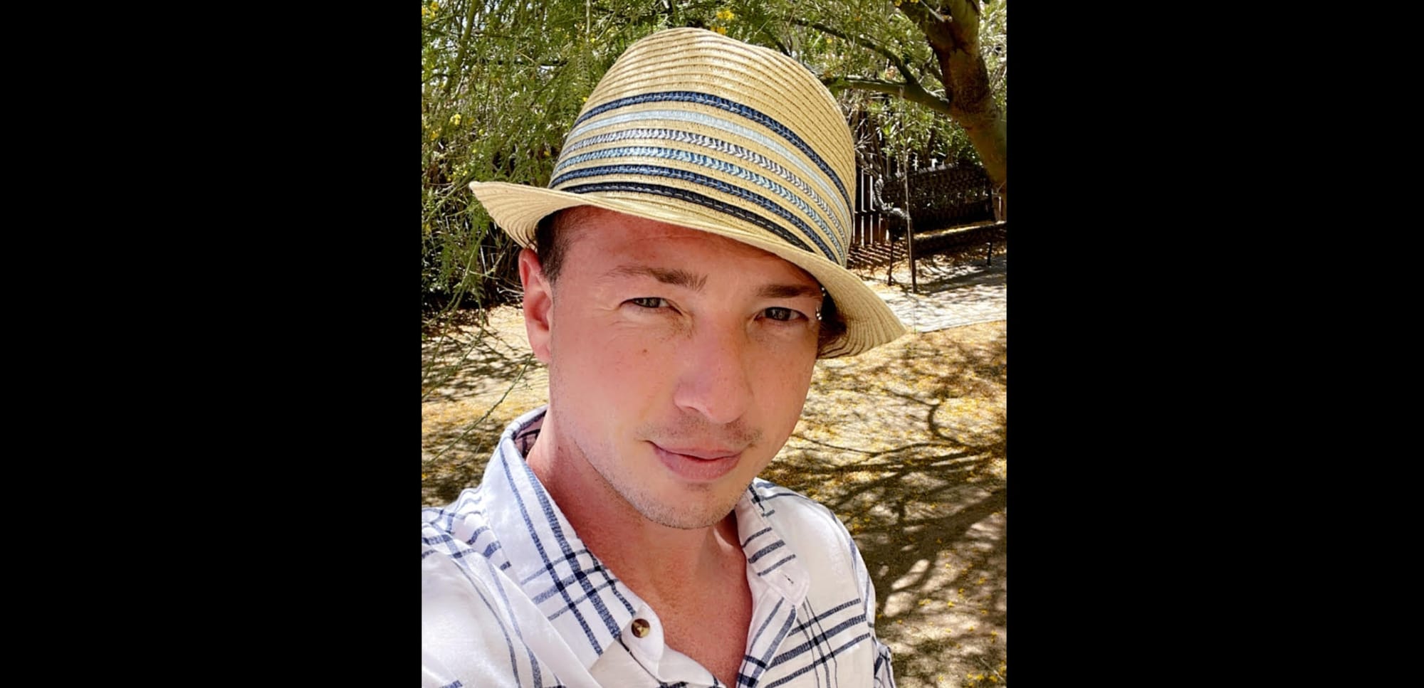  Kaden Vitellaro is wearing a straw hat and checkered shirt, standing outdoors with trees in the background.