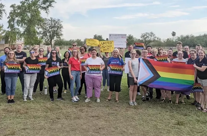 A group of people is gathered outdoors. They're holding signs and a pride flag, showing support against hate.